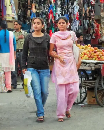 Two young women in salwar suit and jeans walking arm in arm past a fruit cart and shoe stalls