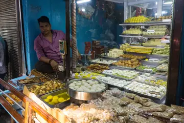 A sweets seller sitting behind trays of sweets and fried snacks at his shop in Varanasi