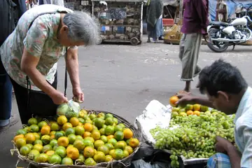 A woman bending to pick oranges from a basket while a seller sorts green grapes beside it at a morning market in Kolkata