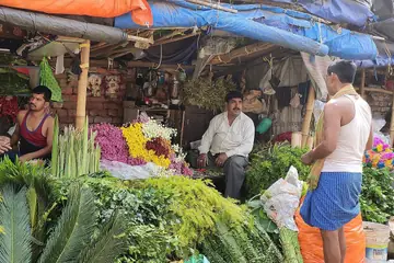 A market stall piled with leafy greens and flowers, with sellers among them under a tarpaulin roof in Howrah