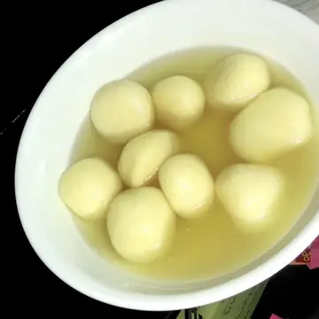 A white bowl of round rasgulla sweets floating in clear sugar syrup