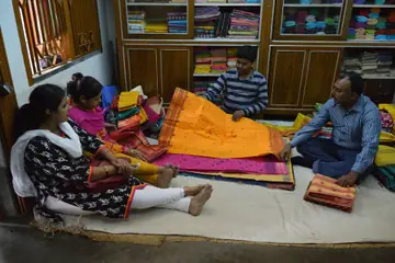 A shopkeeper unfolding an orange tant sari on the shop floor for two women customers, folded saris beside them
