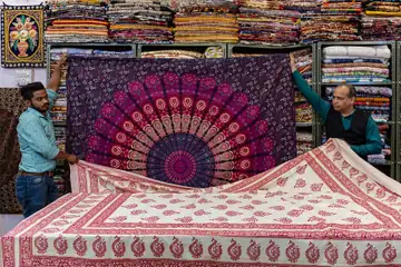 Two shopkeepers holding up and unfolding printed cotton bedspreads in front of shelves of folded cloth in Jaipur