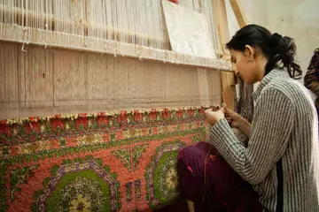 A woman weaving a patterned rug on a tall loom
