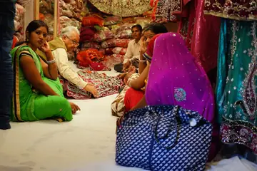 Women customers sitting on a shop floor choosing embroidered cloth with a shopkeeper, saris hanging behind