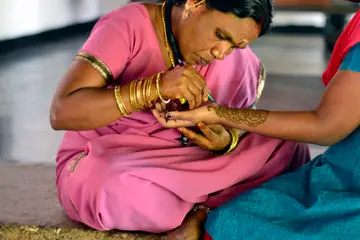 A woman in a pink sari drawing a mehendi pattern on a customer's hand