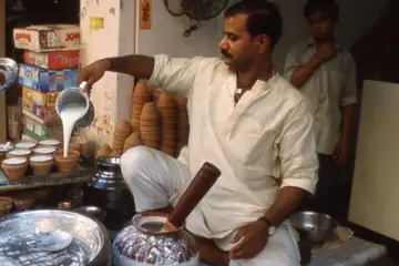 A lassi seller pouring milk into rows of clay cups while a helper serves from a steel tray