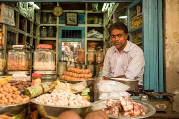 मेवे और मिठाई की ट्रे के पीछे काउंटर पर बैठा दुकान का मालिक, आसपास मर्तबान और शेल्फ़