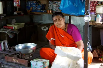 नारंगी साड़ी में एक दुकानदार औरत अपने तराज़ू, एक बोरी और पैकेटों की शेल्फ़ के बग़ल में बैठी है
