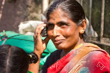 A smiling woman in a red sari holding a mobile phone to her ear at a market