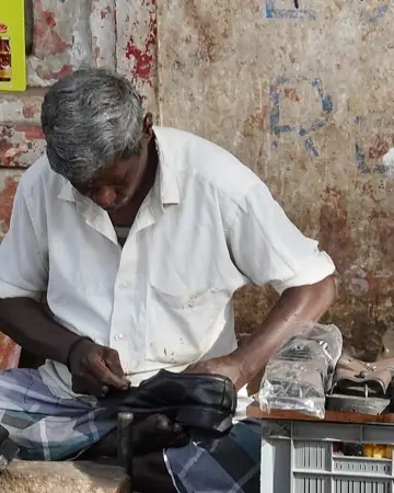 തമിഴ്നാട്ടിലെ തിരുച്ചിറപ്പള്ളിയിൽ വഴിയരികിൽ ഒരു കറുത്ത ഷൂ തുന്നുന്ന ചെരുപ്പുകുത്തി, അരികിൽ നന്നാക്കിയ ഷൂസ്
