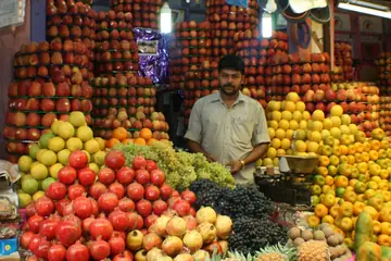 A fruit seller standing behind tall heaps of apples, pomegranates, oranges and grapes with a scale at his counter