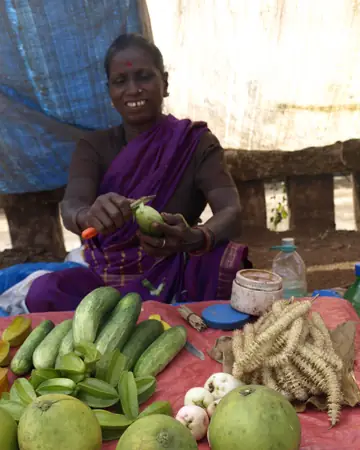 A smiling woman vegetable seller peeling a vegetable, with cucumbers and gourds in front of her