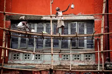 Two painters working on a bamboo scaffold in front of a red-orange building, one reaching up with a paint bucket in Kolkata