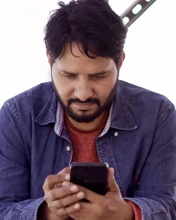 A man in a denim shirt sitting outdoors and looking down at the phone in his hands
