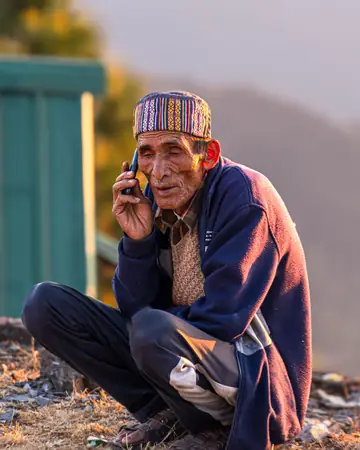 An older man in a patterned cap crouching outdoors with a phone to his ear.