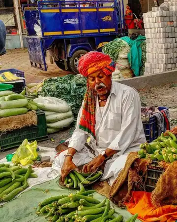 A vegetable seller in a bright orange turban sitting among piles of green gourds and beans
