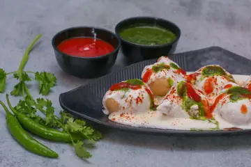 Dahi vada with red and green chutneys and green chillies on a black plate