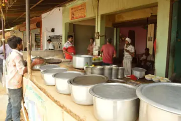 കൗണ്ടറിൽ വലിയ സ്റ്റീൽ പാത്രങ്ങളുള്ള റോഡരികിലെ ധാബ, പിന്നിൽ പാചകക്കാരും കസ്റ്റമർമാരും