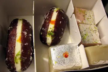 A cardboard box of mithai with a silver-leaf barfi, pistachio slabs and two cut sweets