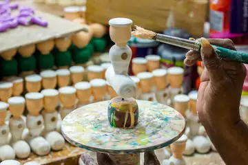 A hand painting a white wooden toy on a turntable at Channapatna, with rows of toy parts behind