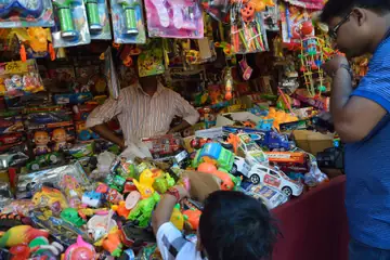 പശ്ചിമ ബംഗാളിലെ കൊൽക്കത്തയിൽ കളിപ്പാട്ട കാറുകളും രൂപങ്ങളും കൂട്ടിയിട്ട കൗണ്ടറിന് പിന്നിൽ നിൽക്കുന്ന കളിപ്പാട്ട വിൽപ്പനക്കാരൻ, ഒരു കസ്റ്റമർ നോക്കുന്നു